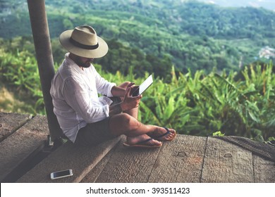 Man traveler is using digital tablet, while is sitting against beautiful Asian scenery during summer journey. Male wanderer is holding touch pad, while is relaxing outdoors during his trip in Thailand