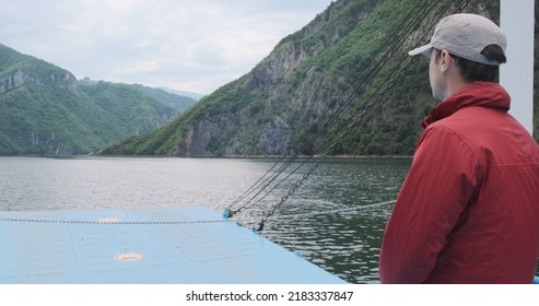 Man Traveling By Ferry Komani Lake In Summer Mountains. Unrecognizable Traveler In Red Jacket On Moving Ship In Albania Alps, Backview. Travel Destination. Adventure At Beautiful Mountains