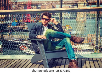 Man traveling in New York. Dressing in black leather jacket, blue jeans, brown boot shoes, wearing sunglasses, a young guy with beard, sitting on chair on deck, relaxing. Boats, bridge on background. 