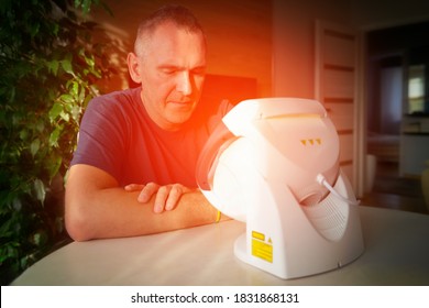 Man Treating Pain In The Sinuses And Head With Infrared Light Therapy. The Man Sits At The Table Near The Healing Lamp.