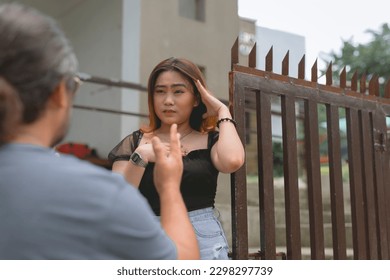 A man tries to reason with his upset younger girlfriend in a black blouse standing beside the gate of her home. - Shutterstock ID 2298297739