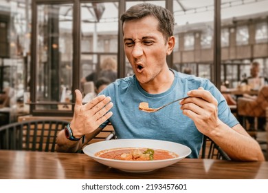 A man tries a spicy and hot red soup in a restaurant and reacts funny emotionally. Seasonings in the national cuisine and an unhealthy diet with overabundance of pepper - Shutterstock ID 2193457601