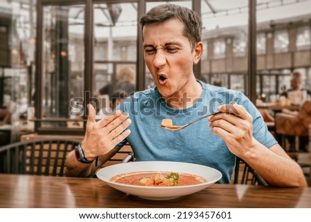 A man tries a spicy and hot red soup in a restaurant and reacts funny emotionally. Seasonings in the national cuisine and an unhealthy diet with overabundance of pepper