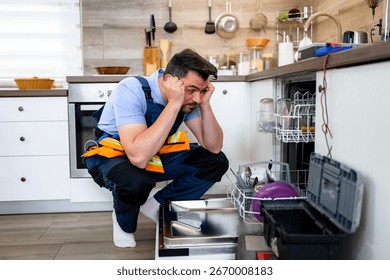 A man is troubleshooting a broken dishwasher while squatting in a well-equipped kitchen.