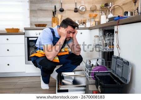 A man is troubleshooting a broken dishwasher while squatting in a well-equipped kitchen.