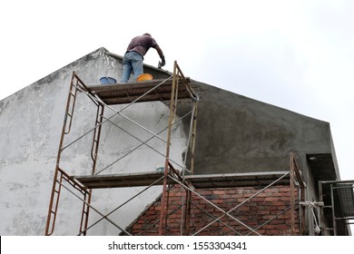 A Man With Trowel Standing High Up On A Makeshift Scaffold Tower, Applying Cement Plaster On The Rooftop Wall As The Worker Rebuild An Exterior Section Of A House Under Renovation And Construction.