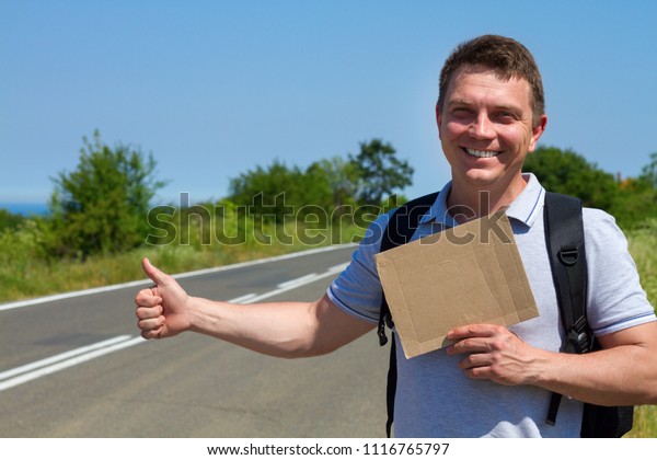 man try to stop car\
with cardboard sign and thumb up gesture, copy space. Man\
travelling by hitchhiking with road on background sea . Travelling\
and hitchhiking concept.
