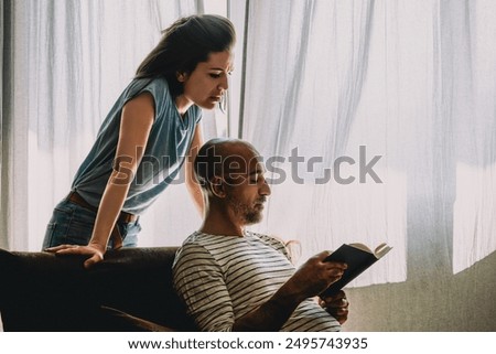 Man trying to avoid a confrontation with his angry wife by concentrating on reading a book, while she is trying to get his attention