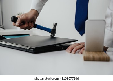 The Man Is Trying To Open A Closed Laptop Using A Hammer. The Businessman Uses A Skeleton Key To Open The Laptop. The Concept Of A Blocked Computer, No Access To Data.