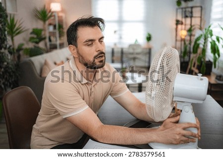 Man trying to refresh himself with a fan during a heat wave at home