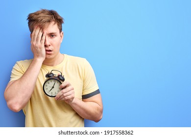 man in t-shirt hold alarm clock in the morning, has no enough sleep, can't wake up. isolated on blue colour background studio portrait.