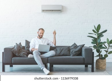 Man Turning On Air Conditioner With Remote Control While Using Laptop On Sofa