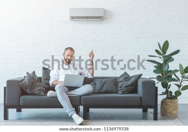 man turning on air conditioner with remote\
control while using laptop on\
sofa