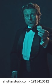 Man In Tuxedo And Bow Tie Holding Casino Dealer Coin. Lit By Blue And Orange Light In Front Of A Dark Wall.