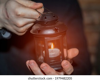 A man in two dirty, working hands holds a candle in a closed candlestick. The concept of a heating point in a shelter, helping the homeless and the poor.