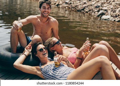 Man With Two Woman Friends Sitting On A Floating Dock In A Lake Drinking Beer. Friends Having Fun At A Lake Relaxing With Drinks In Hand.