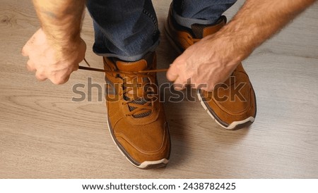 A man is tying the laces on his brown shoes. He is wearing blue jeans, reflecting a casual and modern style. Emphasis on the shoe