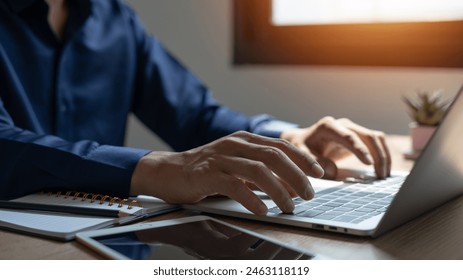 A man is typing on a laptop in front of a window. The laptop is open and the man's hands are on the keyboard. There are several notebooks and a tablet on the desk - Powered by Shutterstock