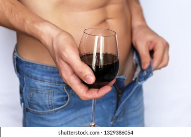 Man In Unbuttoned Jeans With Naked Torso Standing With Glass Of Red Wine In Hands. Concept Of Relaxation, The Negative Effects Of Alcohol Consumption, Impotence, Romantic Sex Date