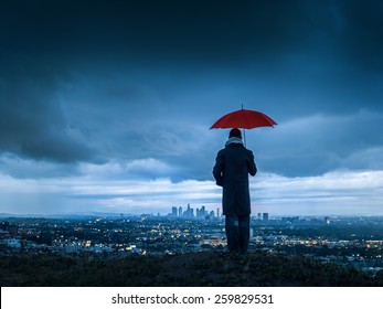 Man under red umbrella overlooking stormy Los Angeles cityscape from Hollywood Hills at twilight.  - Powered by Shutterstock