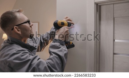 Man in uniform and glasses screws security camera mount into concrete wall with portable screwdriver. Installer or repairman works in apartment. Installation of CCTV cameras. Repair work.