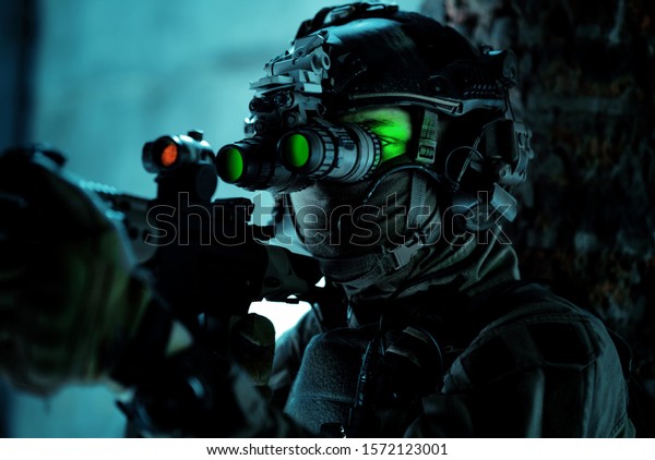 Man in uniform with machine gun and\
turned on night vision device beside brick wall. Closeup airsoft\
soldier with green light on face in night\
building