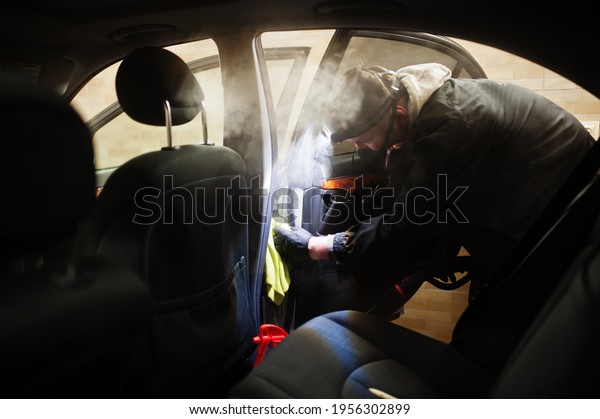 Man in uniform and respirator, worker of car\
wash center, cleaning car interior with hot steam cleaner. Car\
detailing concept.