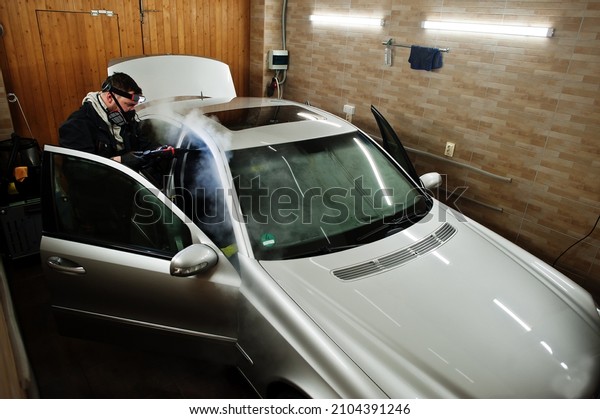 Man in uniform and respirator, worker of car\
wash center, cleaning car interior with hot steam cleaner. Car\
detailing concept.