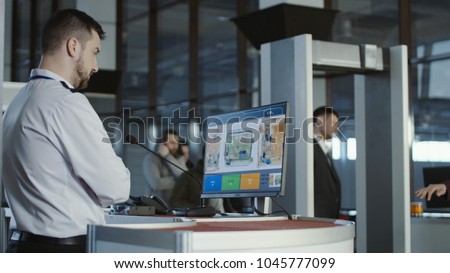 Man in uniform standing at counter at checking point and watching at monitor with x-ray of luggage.