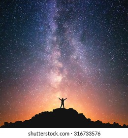 Man and the Universe. A person is standing on the top of the hill next to the Milky Way galaxy with his hands raised to the air. 