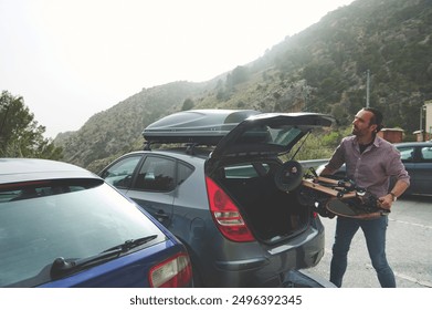 A man unloads a longboard from a car trunk, ready for an adventure amidst picturesque mountains. Capturing travel, leisure, and outdoor activities. - Powered by Shutterstock