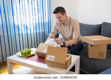 Man Unpacking Awaited Parcel, Looking Inside, Sitting At Sofa, Satisfied Happy Customer Opening Cardboard Box With Online Store Order, Good Shipping Delivery Service. 