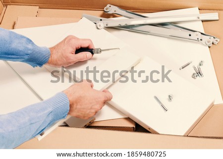 Man unpacks and assembles flat packed furniture isolated. Box with white diy funiture and hands with a screwdriver and screws. Relocation or moving into new house concept with copy space
