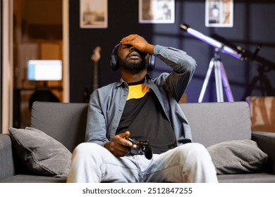 Man upset after losing while playing videogames on console system with joypad in apartment late at night. Irked gamer frustrated with defeat during multiplayer game, sulking on couch