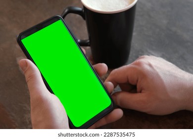 Man Use of Mobile Phone with Blank Green Screen in a Cafe on Rustic Vintage Wooden Table. Male Holding Smart Phone Empty ChromaKey Screen Mock up in Coffee Shop. Morning. Showing Mock up. - Powered by Shutterstock