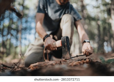 A man uses an axe to split a log while kneeling on the forest floor. The outdoor camping scene shows hands-on wood chopping next to fallen branches and dry leaves.
