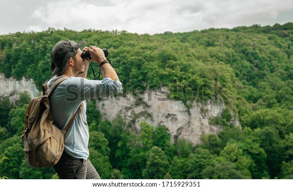 A man uses binoculars while standing at\
a height with a backpack on his back inspecting the forest.\
Environmental studies and forest fire\
protection.