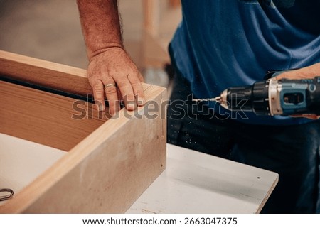 A man uses a cordless drill to insert screws into a cabinet made of MDF and wood in a workshop setting, focusing on detailed and precise furniture assembly