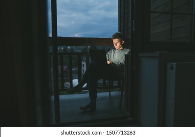 Man Uses A Laptop At Night On A Balcony In An Apartment,sits In A Chair,looks At A Computer Screen With A Serious Face.Freelancer Works On Laptop At Home On A Balcony In A Country House Late At Night