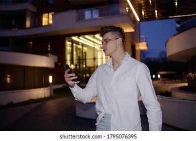 The Man Uses The Phone At Night. A Teenage Man At Night With A Phone Goes On A Date, Acquaintance On The Internet With A Rich Guy. Young Man. Glasses