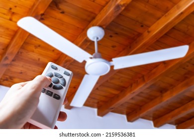 A man uses a remote control to turn on a white ceiling fan mounted in a house with wooden ceilings.	