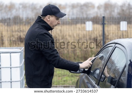 Man uses a scraper to clean the side windows of his car in an outdoor space.