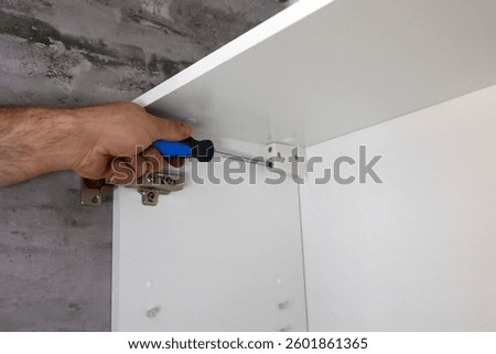 A man uses a screwdriver to fix a bracket mounted on a white rack, a cabinet mounted on the wall. This home improvement task is performed in a modern interior. 
