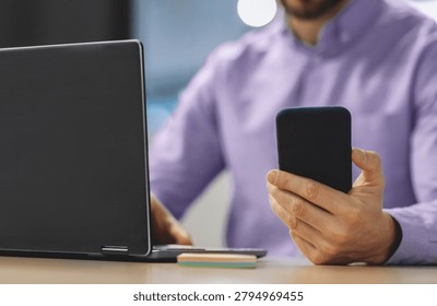 Man Uses Smartphone While Working on Laptop at a Desk During the Day