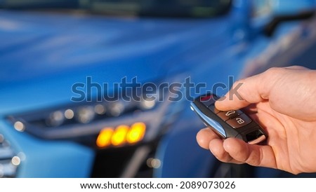 Man uses thumb to press black button on remote control key removing alarm signalling from bright blue car and headlights flash closeup