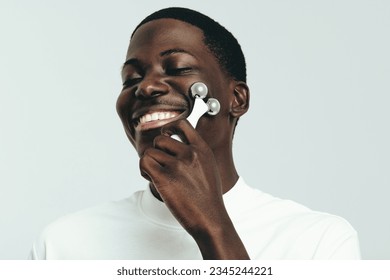 Man using a 3D face roller to massage his skin, working out any tension and promoting a healthy glow. Happy young pampering his skin with a beauty tool as he indulges in his daily skincare routine. - Powered by Shutterstock