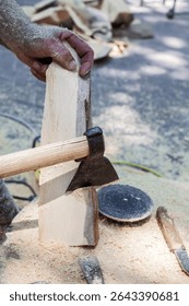 Man is using an axe to cut a piece of wood. The scene is set in a wooded area, with the man standing in front of a pile of wood. The atmosphere of the image is calm and focused