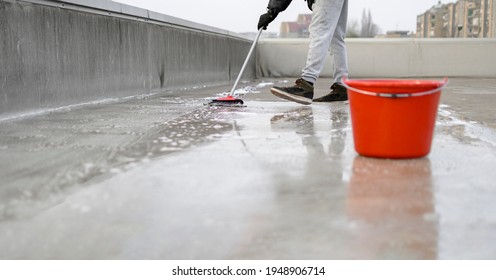 Man Using Broom And Water To Clean Dirt From The Rooftop. Red Plastic Bin On The Right Side Of Photography. He Is Wearing A Black Jacket, Gloves, And Gray Sweat Suit. Before And After Situation.