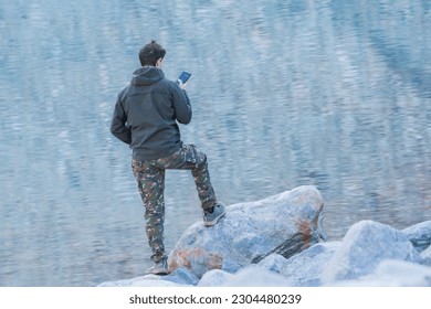 Man using a cell phone in front of a mountain lake. Photo from behind