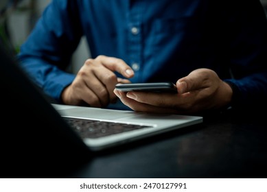 A man is using a cell phone while sitting at a desk. Concept of productivity and focus, as the man is engaged in a task on his phone. The laptop on the desk suggests that he may be working or studying - Powered by Shutterstock